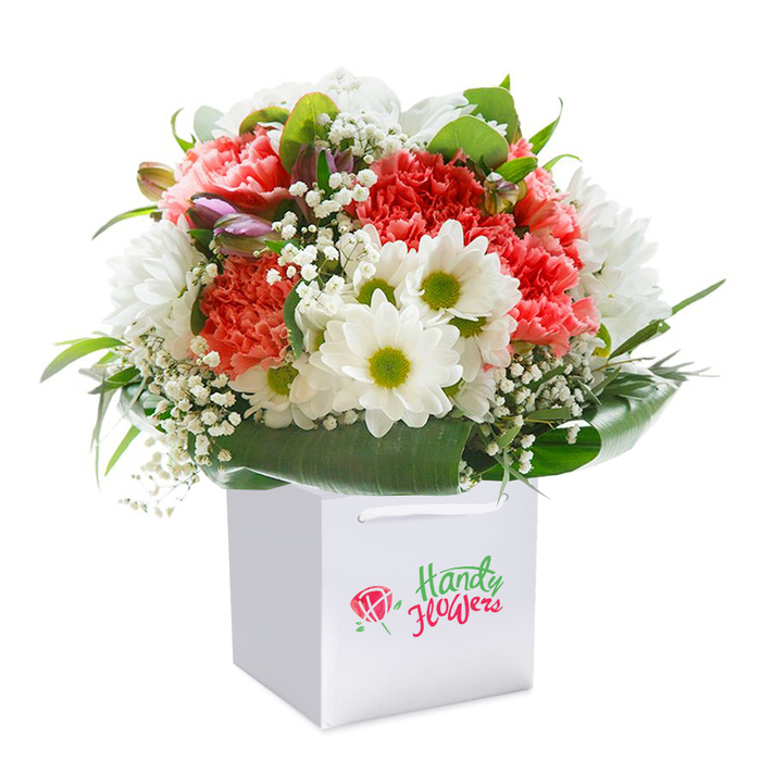 Expertly crafted by a florist in Hither Green, this lush and uplifting bouquet stands upright in a clean, contemporary white square gift bag, set before a seamless white background that enhances every petal and flourish. The arrangement brims with plush coral and peach carnations, their ruffled petals layered in abundance to create texture and vibrancy. Scattered throughout are crisp, sparkling white daisies with lime-green centers, offering a nod to the cheerful wildflowers that bloom along local garden paths. Tiny clusters of ethereal baby's breath provide delicacy, filling the spaces between blooms like little pockets of light. Hints of unopened flowers with soft purple tips peek out from the left, adding depth and anticipation much like the budding trees along Meridian South in spring. The bouquet's perimeter is gracefully encircled with deep green foliage, including broad, shiny leaves that cradle the blooms and add a dramatic, structural finish. With its perfectly balanced, rounded form and elegant white ribbon handles, the presentation is ready for gifting-a lovely choice for an anniversary near Hither Green station, a birthday surprise in nearby Catford, or an expression of gratitude for local friends and family. The result is a bouquet that reflects both the freshness of the blooms and the genuine care of local floristry, making it a standout gesture for any special occasion.