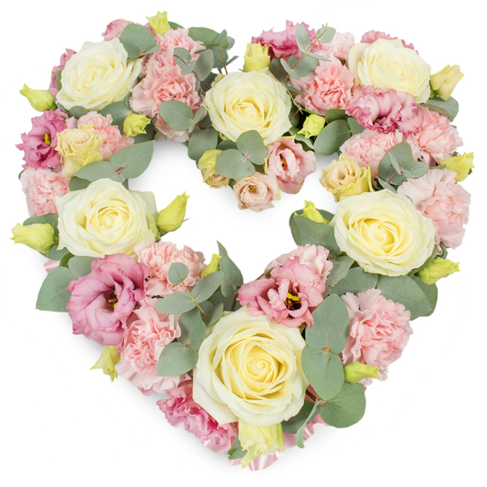 In this image, a generously filled heart-shaped funeral wreath appears almost to float against a bright white background, every part of its shape covered with soft flowers and foliage. The heart is clearly defined yet gently rounded, created from a careful balance of creamy yellow and ivory roses, blush-toned carnations, and deeper pink lisianthus. The roses are the most prominent, their velvety petals tightly spiralling at the centre and then broadening outward, giving each bloom a classic, timeless elegance that often comforts families in Hither Green saying goodbye to someone special. Nestled around and between them are clusters of pastel pink carnations, each head made up of many frilled petals that ruffle into one another, lending a sense of tenderness and cushioning to the overall design. Threads of richer colour wind through the wreath in the form of magenta and mauve lisianthus, their softer, more open petals curling at the edges and, in some blooms, revealing slightly darker centres that draw the eye inward. Silvery-green eucalyptus leaves run throughout the arrangement, their rounded, slightly waxy leaves adding subtle structure, cool contrast, and a gentle, aromatic freshness reminiscent of the calm air in nearby Manor House Gardens. Tiny yellow-green buds dot the composition, suggesting life still unfolding even in a time of parting. Every stem has been placed close to its neighbours so the heart appears full and luxurious, with layers of texture and colour that invite a lingering gaze. This kind of floral tribute is ideal for funerals, memorials, or anniversaries within the Hither Green community, a quiet yet eloquent symbol of enduring love and heartfelt sympathy.