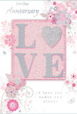 An anniversary card with a soft, romantic design rests against a pale surface, its artwork evoking the tender calm of an early evening walk through Hither Green. The main feature is the word "LOVE," rising from the card in embossed letters coated with delicate blush and pearly glitter, giving a tactile, almost jewel-like sheen. The letters sit on a smooth, dusky pink background that appears lightly mottled, as though painted with a soft brush, lending warmth and depth to the scene. From behind this central word, graceful lines of stylised flowers and foliage curl outward, drifting across the card in airy arcs that resemble the natural sweep of hand-tied stems in a florist's design. The blossoms are drawn in fine white and silver outlines, with some petals gently shaded in blush, mauve-tinged pink, and pale grey, evoking the soft tones of anniversary roses and delicate spring flowers you might see in Hither Green front gardens. Subtle scrolls, dots, and filigree accents fill in the spaces, adding a sense of movement and celebration without overwhelming the eye. The printed message, set in an elegant, flowing script, offers heartfelt anniversary wishes and hints at long years of companionship and shared history. Its understated grey ink complements the silvery details scattered among the blooms. The overall effect is refined yet tender, like a carefully chosen card tucked alongside a bouquet ordered for same-day delivery in Hither Green, capturing both the grace of the occasion and the quiet strength of lasting love.